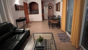 Una sala de estar con una mesa de centro de vidrio, un sofá de cuero negro, un comedor con mesa y sillas, y una cocina con armarios de madera y un refrigerador.