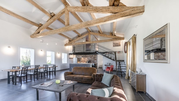 A spacious living area with a high ceiling, exposed wooden beams, a cozy seating area with a sofa and armchairs, a dining table with chairs, and a kitchen in the background. - Loft in Provence in Moustiers Sainte Marie and near the Sainte Croix Lake (Moustiers-Sainte-Marie)