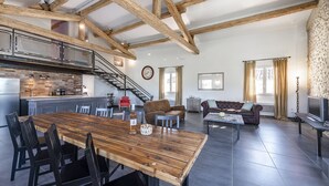 A spacious living area with a wooden dining table, a sofa, and a TV. A kitchen with a modern design and a staircase leading to an upper level is visible. - Loft in Provence in Moustiers Sainte Marie and near the Sainte Croix Lake (Moustiers-Sainte-Marie)