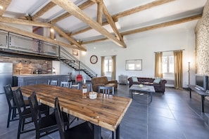 A spacious living area with a wooden dining table, a sofa, and a TV. A kitchen with a modern design and a staircase leading to an upper level is visible.