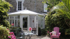 Ein Wintergarten mit Tisch und Stühlen, umgeben von viel Grün und einem Steinhaus.