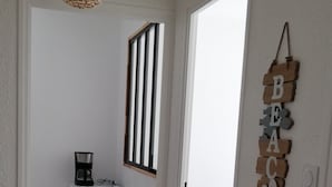 Un pasillo con una cafetera sobre una mesita, una lámpara colgante de mimbre y una obra de arte con palabras fijada en la pared que dice “BEACH”.