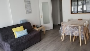 Una sala de estar con un sofá gris oscuro, una mesita de madera y una mesa de comedor con sillas. Una puerta blanca da a otra habitación.