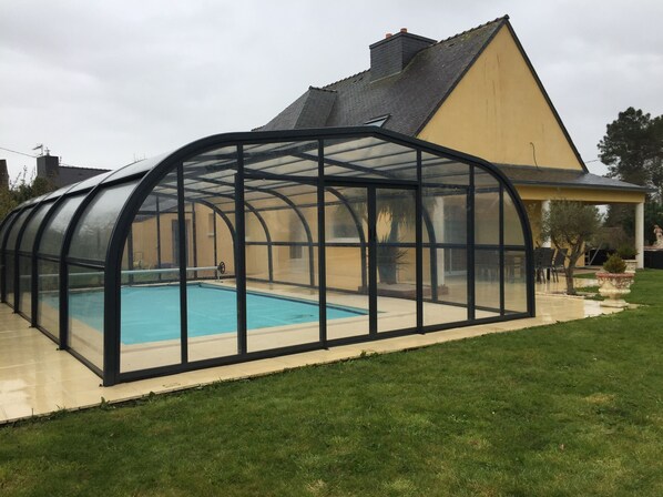 A covered swimming pool with a glass enclosure, surrounded by a lawn and a house with a sloped roof.