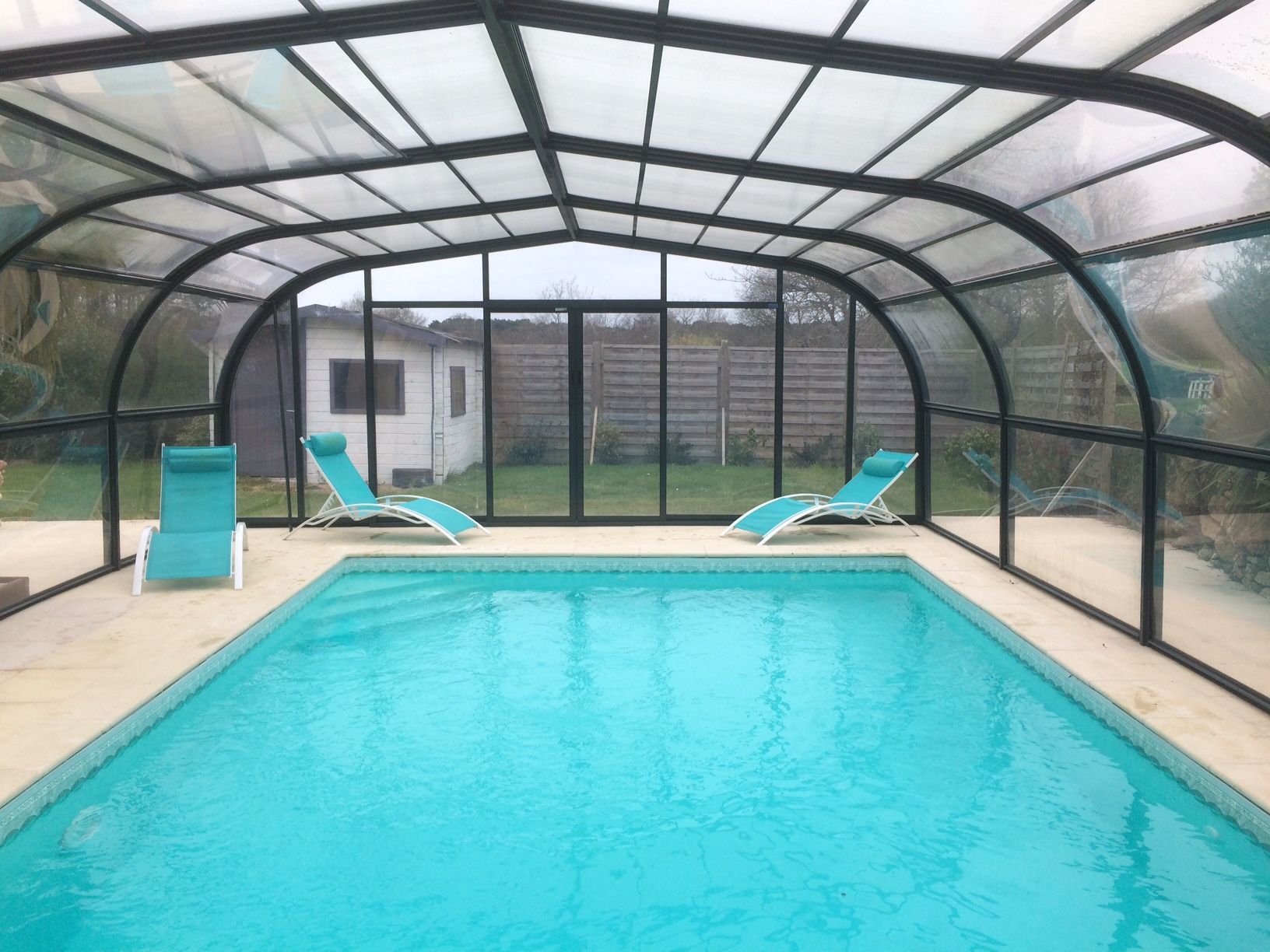 A covered swimming pool with two lounge chairs and a house in the background.