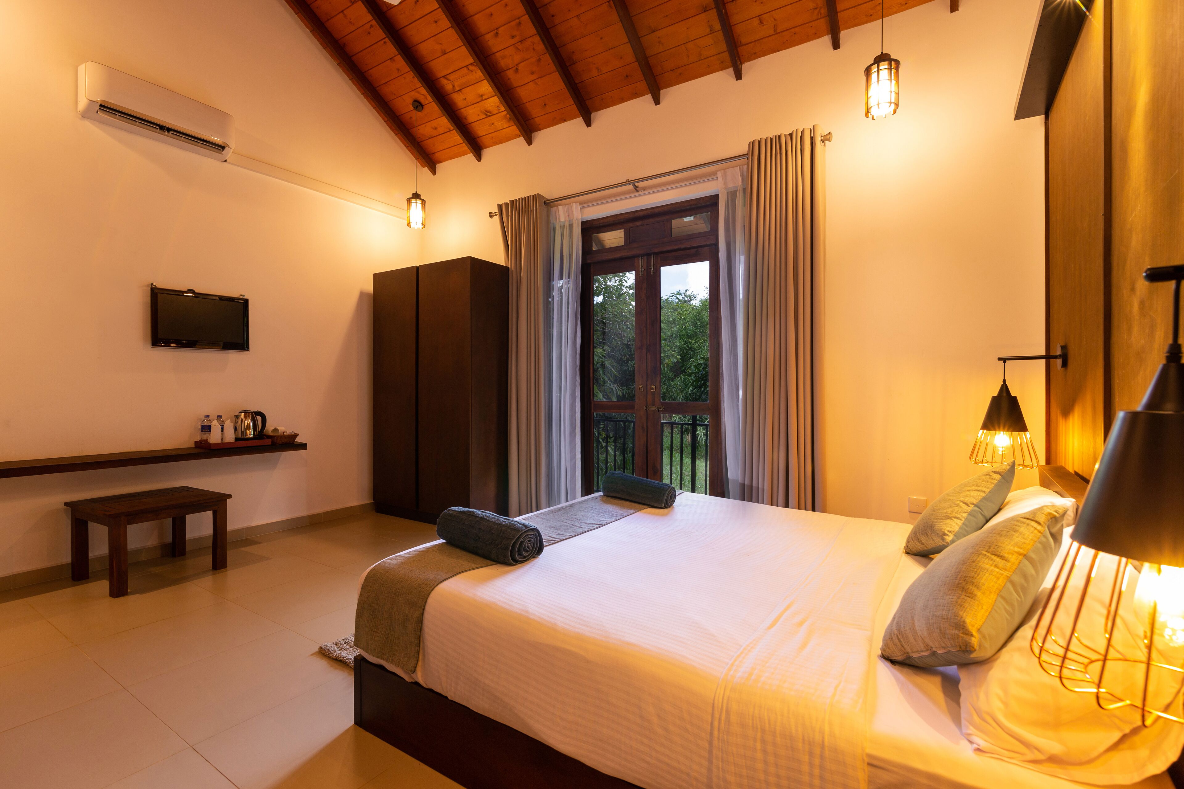 A bedroom with a bed, bedside lamps, a TV, a window with curtains, and a wooden ceiling.