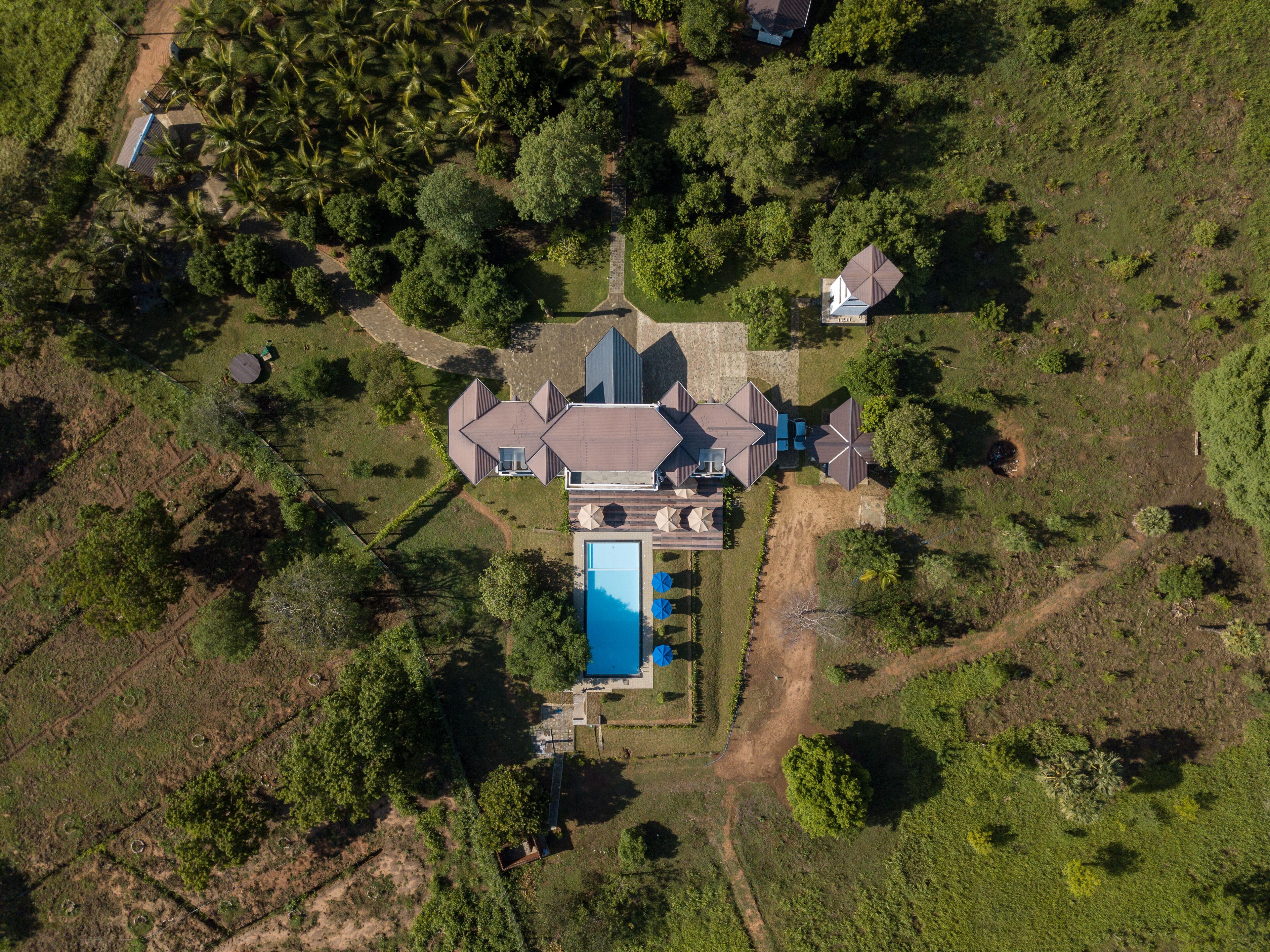 An aerial view of a large, modern house with a swimming pool, surrounded by dense greenery and palm trees.