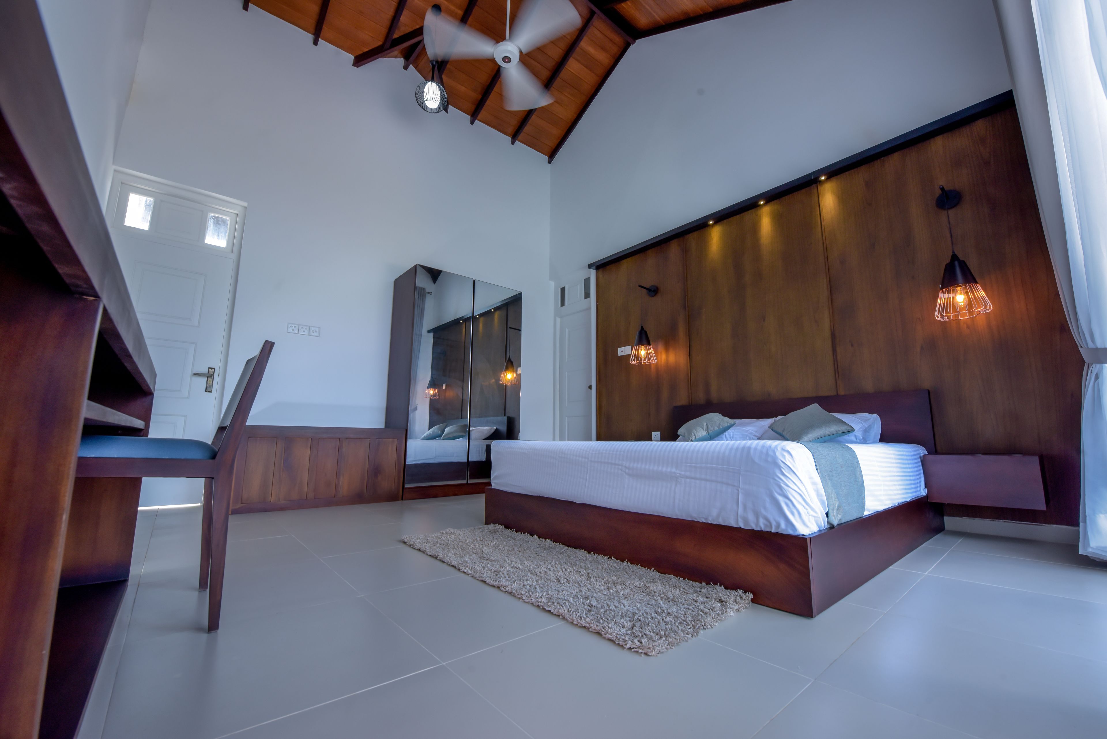 Un dormitorio moderno con una cama grande, cabecera de madera, ventilador de techo e iluminación colgante.