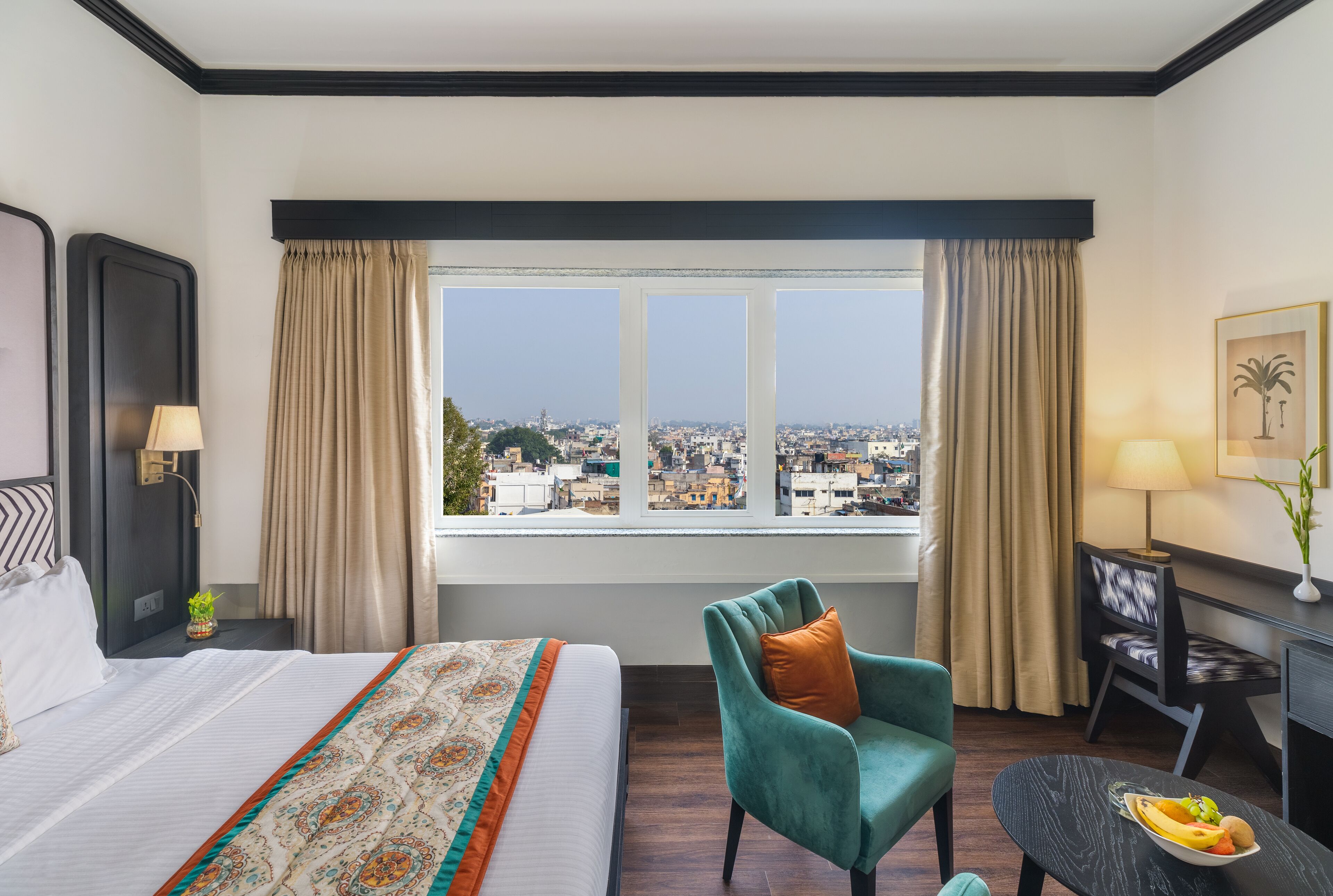 Een hotelkamer met een groot bed, een bureau met stoel, uitzicht op de stad door het raam en een klein tafeltje met fruit.