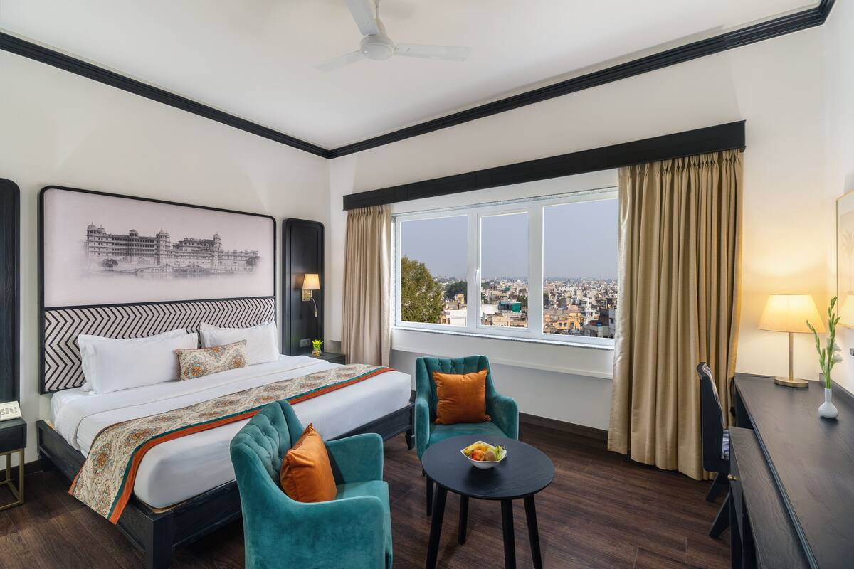 Ein Hotelzimmer mit einem großen Bett, zwei Sessel, einem kleinen Tisch mit Obst, einem Schreibtisch, einer Lampe und Blick auf eine Stadtansicht durch das Fenster.