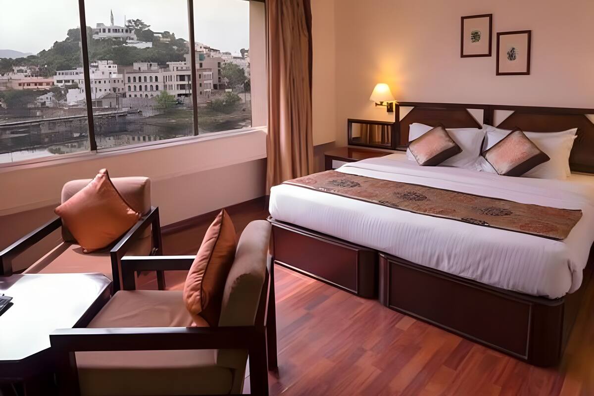 Ein Hotelzimmer mit einem großen Bett, einem Schreibtisch, einem Stuhl, einem Nachttisch und einem Fenster mit Blick auf die Stadt.