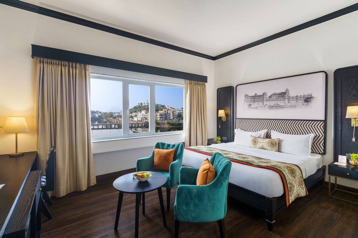 Ein Hotelzimmer mit einem Bett, zwei Stühlen, einem kleinen Tisch, einer Lampe und Blick auf die Stadt durch das Fenster.