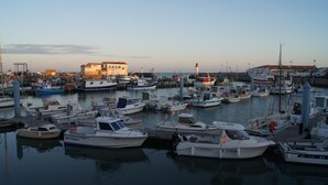 Un port de plaisance où sont amarrés différents bateaux, un bâtiment imposant portant l’inscription « S.N.S.M. », et un phare en arrière-plan.