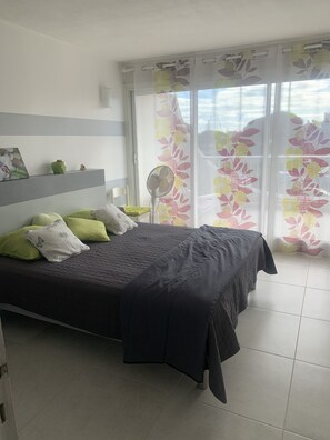 A bedroom with a bed, pillows, a fan, a window with floral curtains, and a view of the sky.