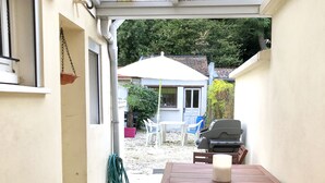 Een overdekt terras met een houten tafel en stoelen, een barbecue en een hangplant.