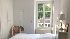 Een slaapkamer met een bed, een stoel, een raam, een radiator en een plank met een lamp en een fotolijst.