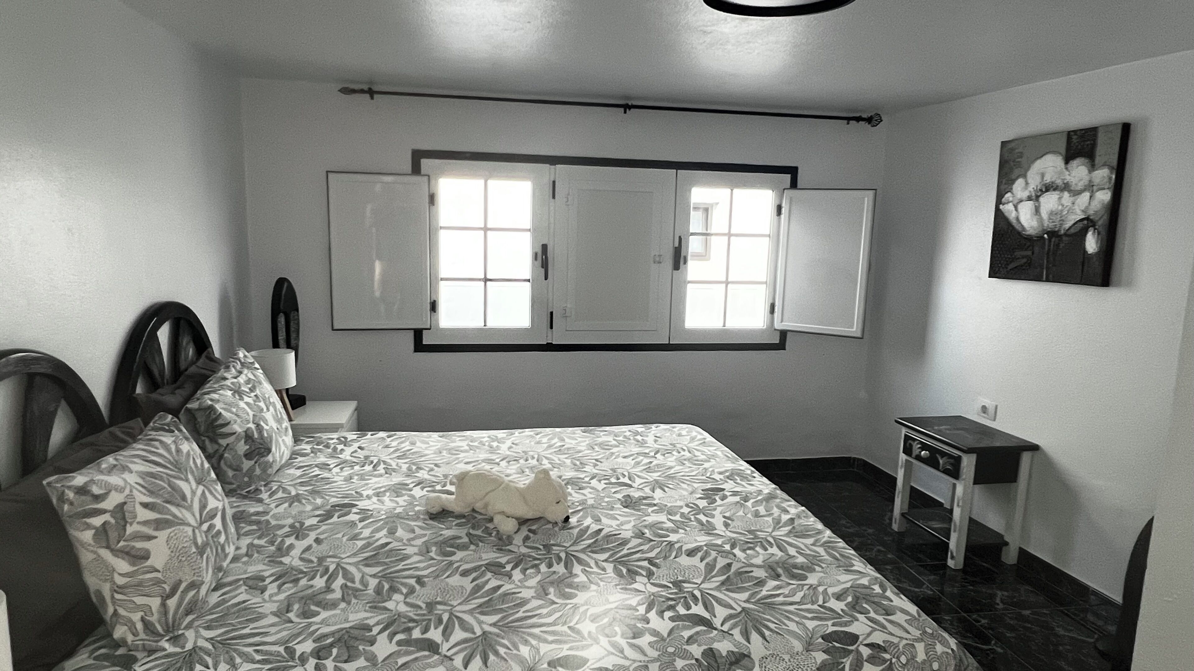 Ein Schlafzimmer mit einem Bett, einem Fenster, einem Nachttisch, einem Sessel, einem kleinen Tisch und einem Bild an der Wand.