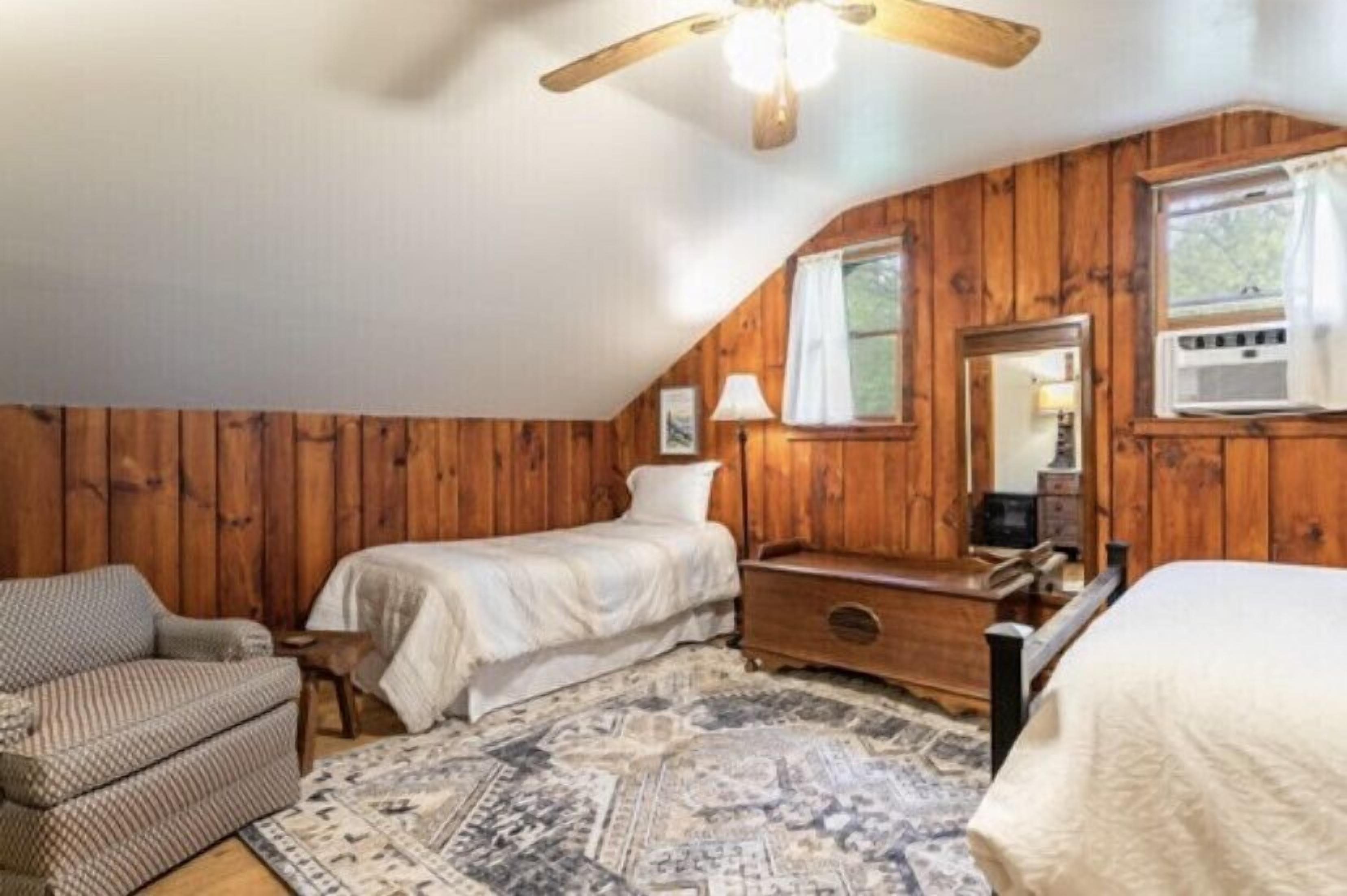 A cozy bedroom with a wooden paneled wall, a single bed, a nightstand, a chair, and a ceiling fan.