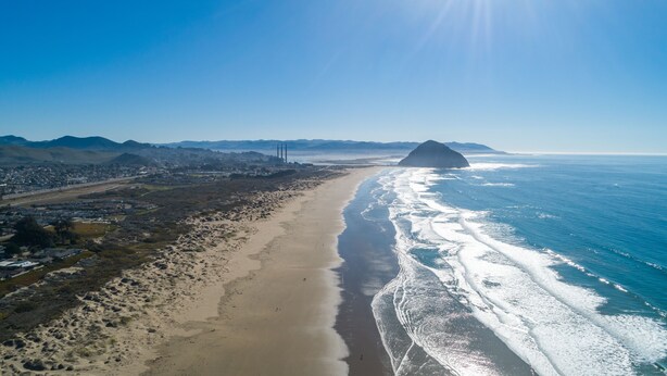 An aerial view of a beach with a long stretch of sand, a clear blue ocean, and distant hills. - Beach House Perfect for your Next Getaway! (Morro Bay)
