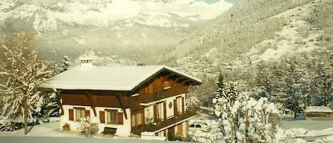A wooden chalet with a snow-covered roof, surrounded by snow-covered trees and mountains in the background.