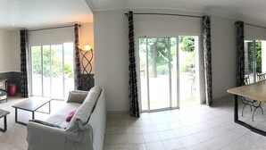 A spacious living area with a sofa, a coffee table, a TV, and a dining table with chairs. There is a balcony with a view of greenery.