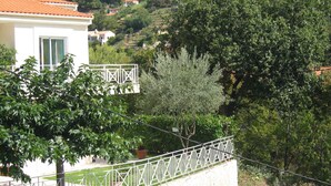 A white building with a red-tiled roof, surrounded by lush greenery and hills in the background.
