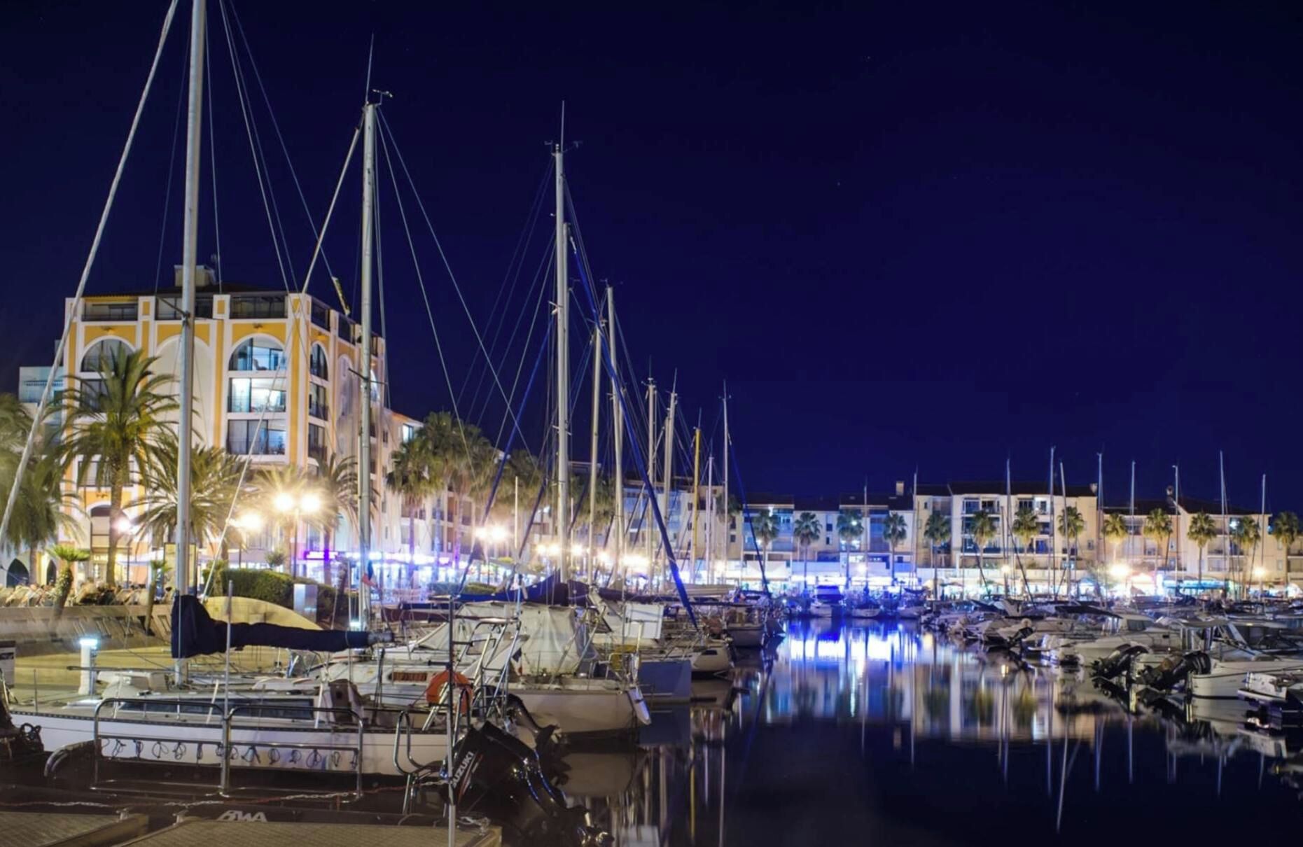 Un puerto deportivo de noche con barcos amarrados, un edificio de varios pisos y palmeras.