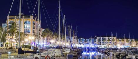 A marina at night with boats docked, a building with multiple floors, and palm trees.