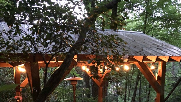 A wooden deck with outdoor furniture, surrounded by trees and a pergola with string lights. - Minutes To Town, Wooded 2 Acre Retreat (Highlands)