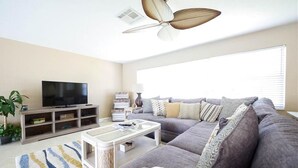En stue med en grå sofa, et glasbord, et tv på et træstel og en loftventilator.