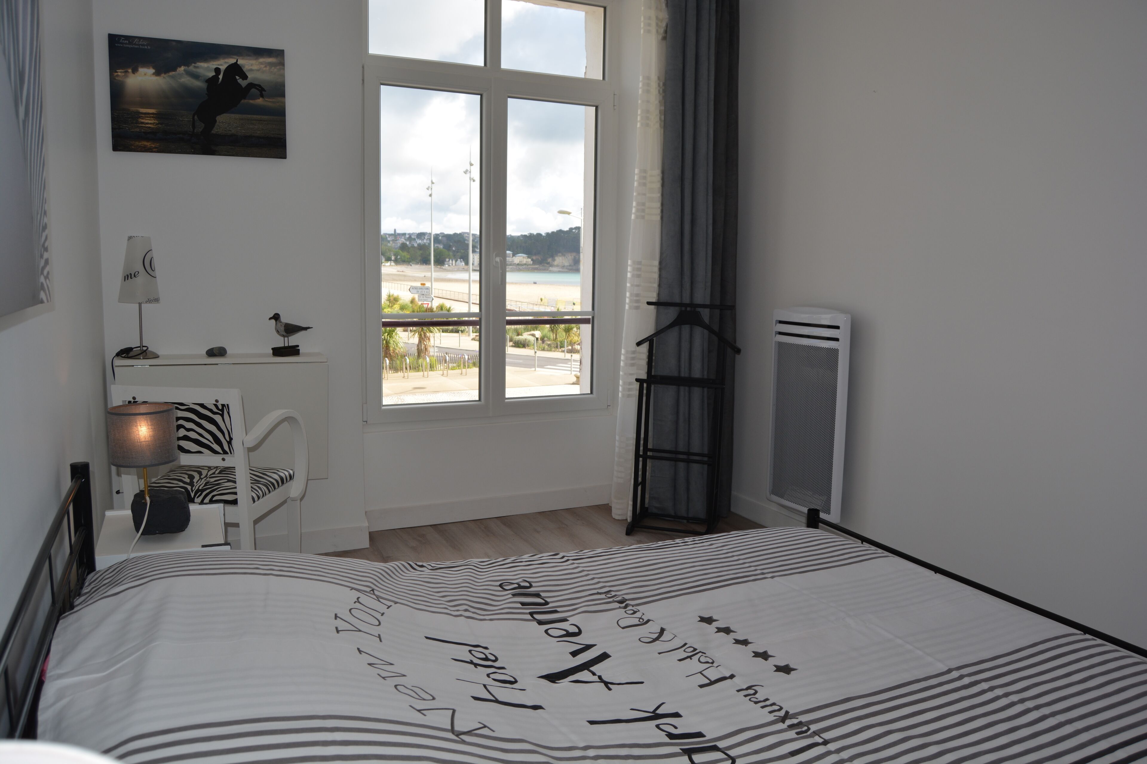 Ein Schlafzimmer mit großem Fenster, einem Bett mit gestreiftem Überwurf, einem zebra-gemusterten Sessel, einer Lampe und einem kleinen Tisch mit Vogel-Figur.