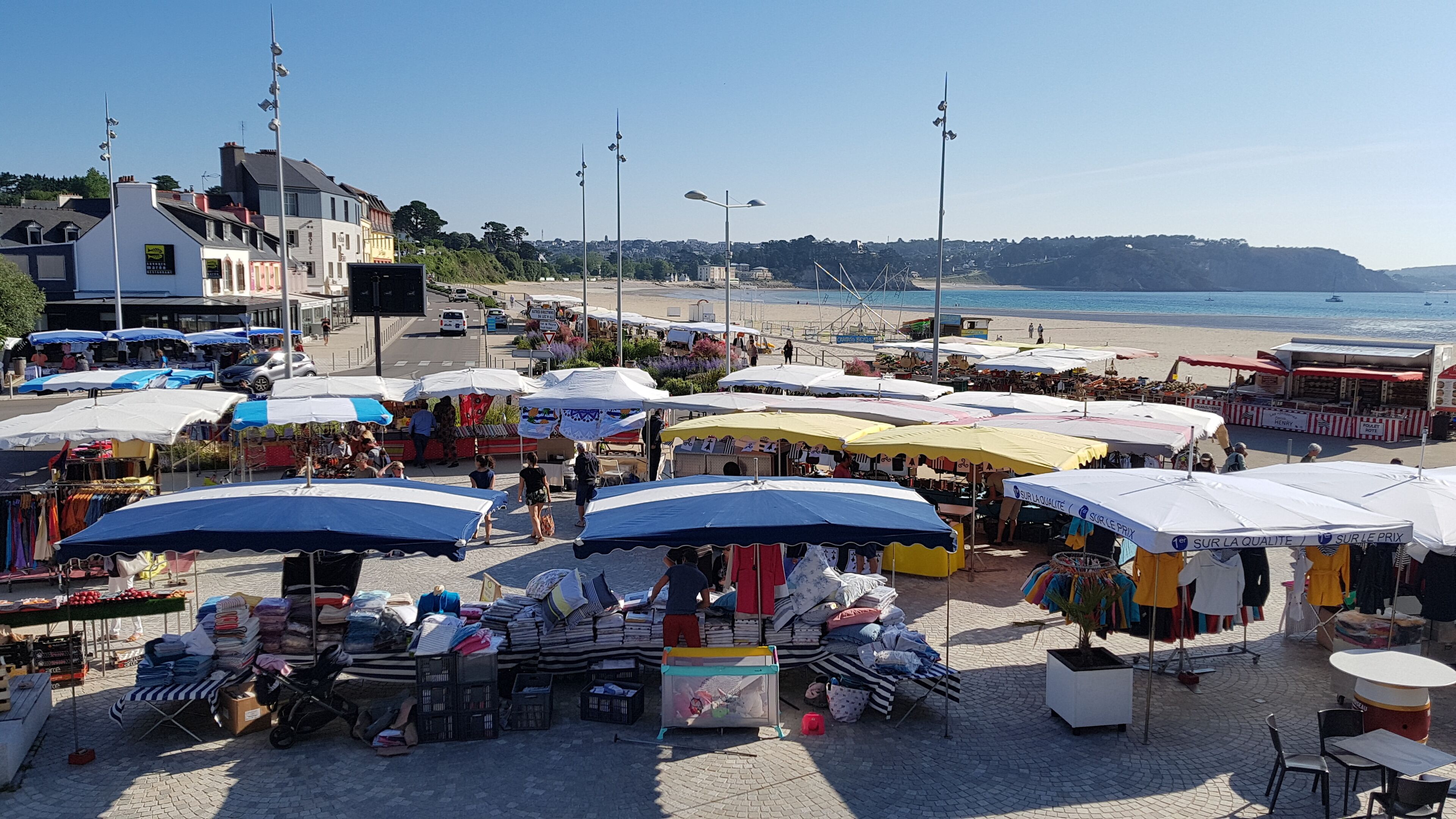 A bustling outdoor market with various stalls and umbrellas, located near a beach and buildings.