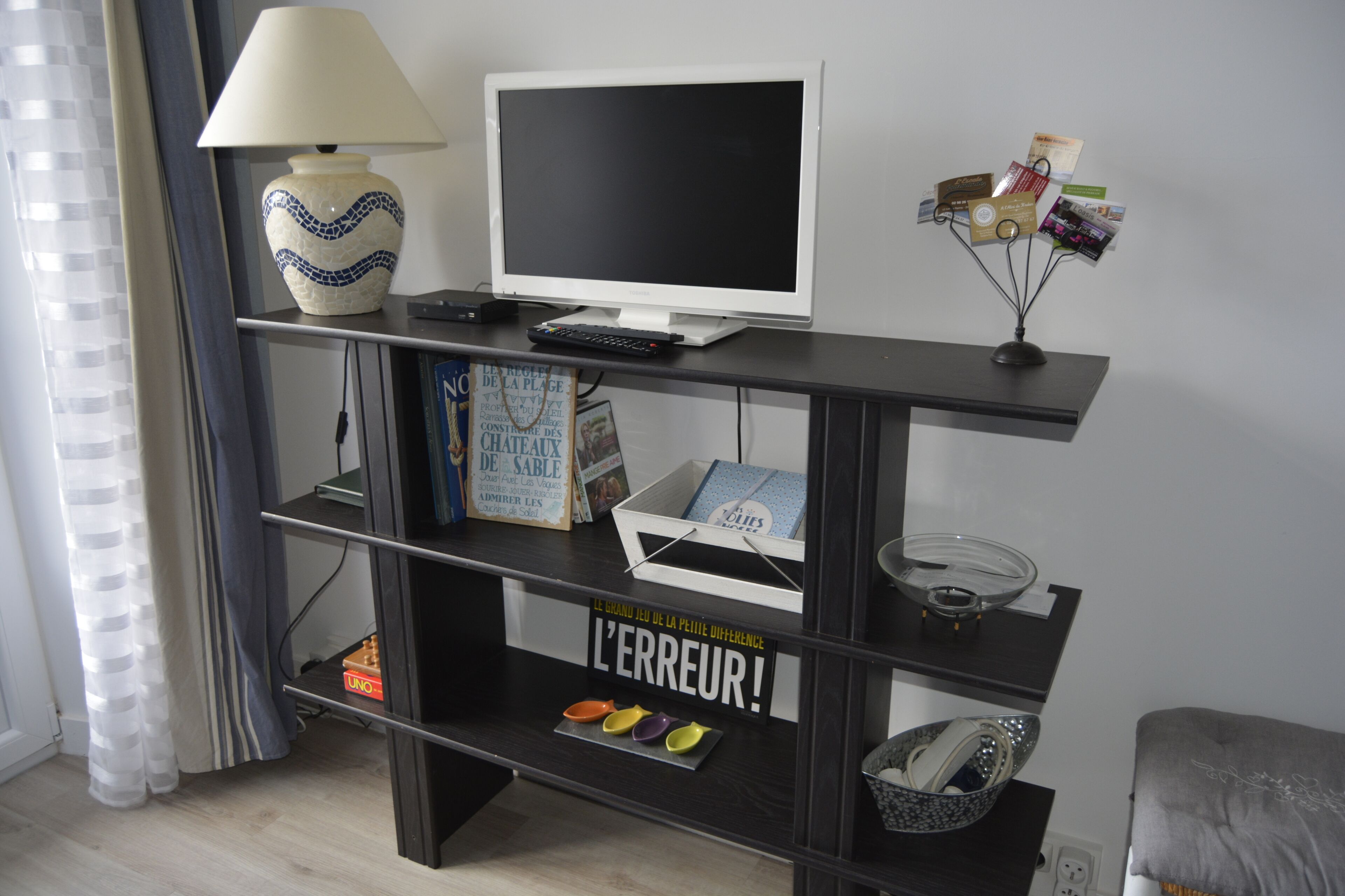 Een moderne woonkamer met een televisie op een zwart voetstuk, een lamp en een plank met boeken en decoratieve objecten.