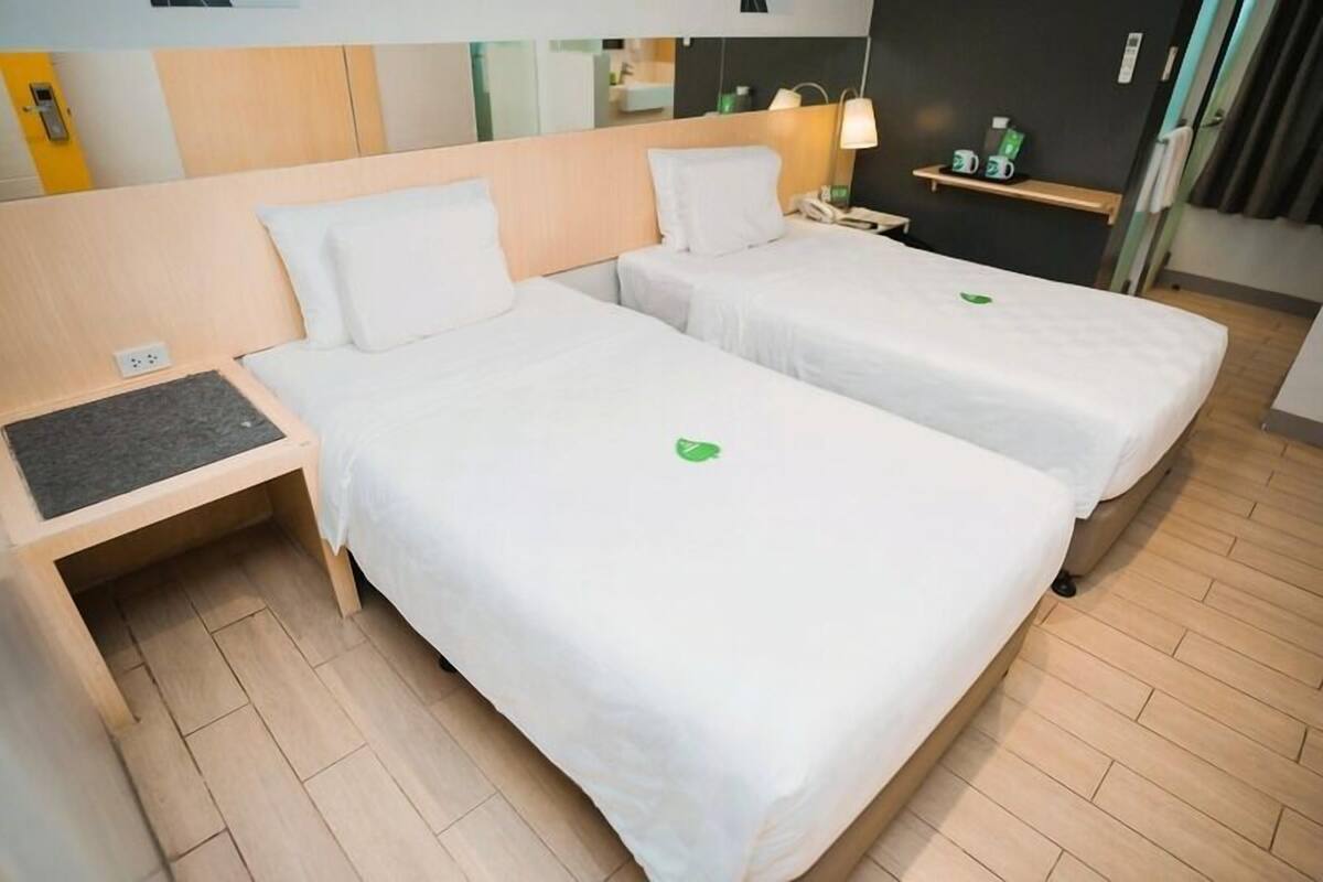A hotel room with two beds, a bedside table, a small shelf with items, and a mirror on the wall.