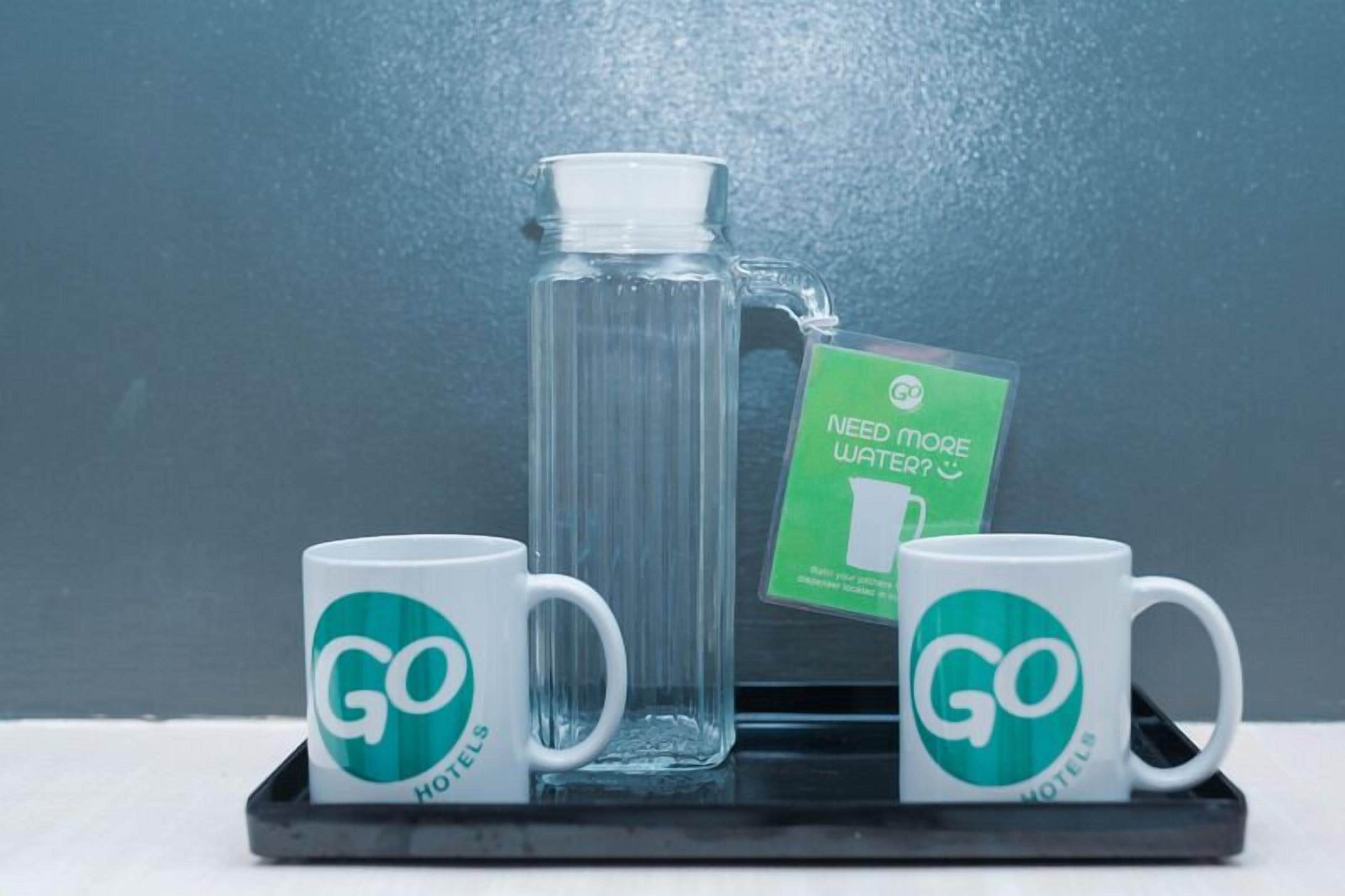 Um jarro de vidro com água, duas canecas brancas com o logo 'GO HOTELS' e um cartão verde com o texto 'PRECISA DE MAIS ÁGUA?' sobre uma bandeja preta.