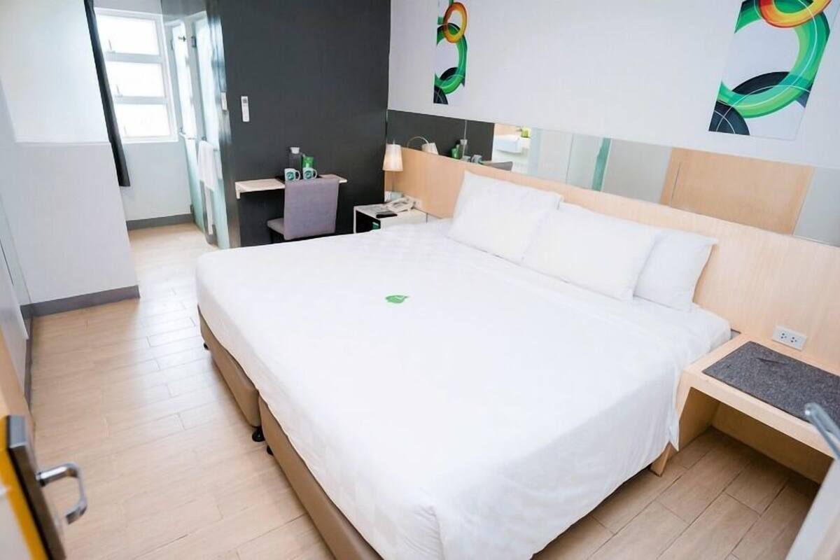 A hotel room with a large bed, a bedside table, a desk with a chair, a mirror, and artwork on the walls.