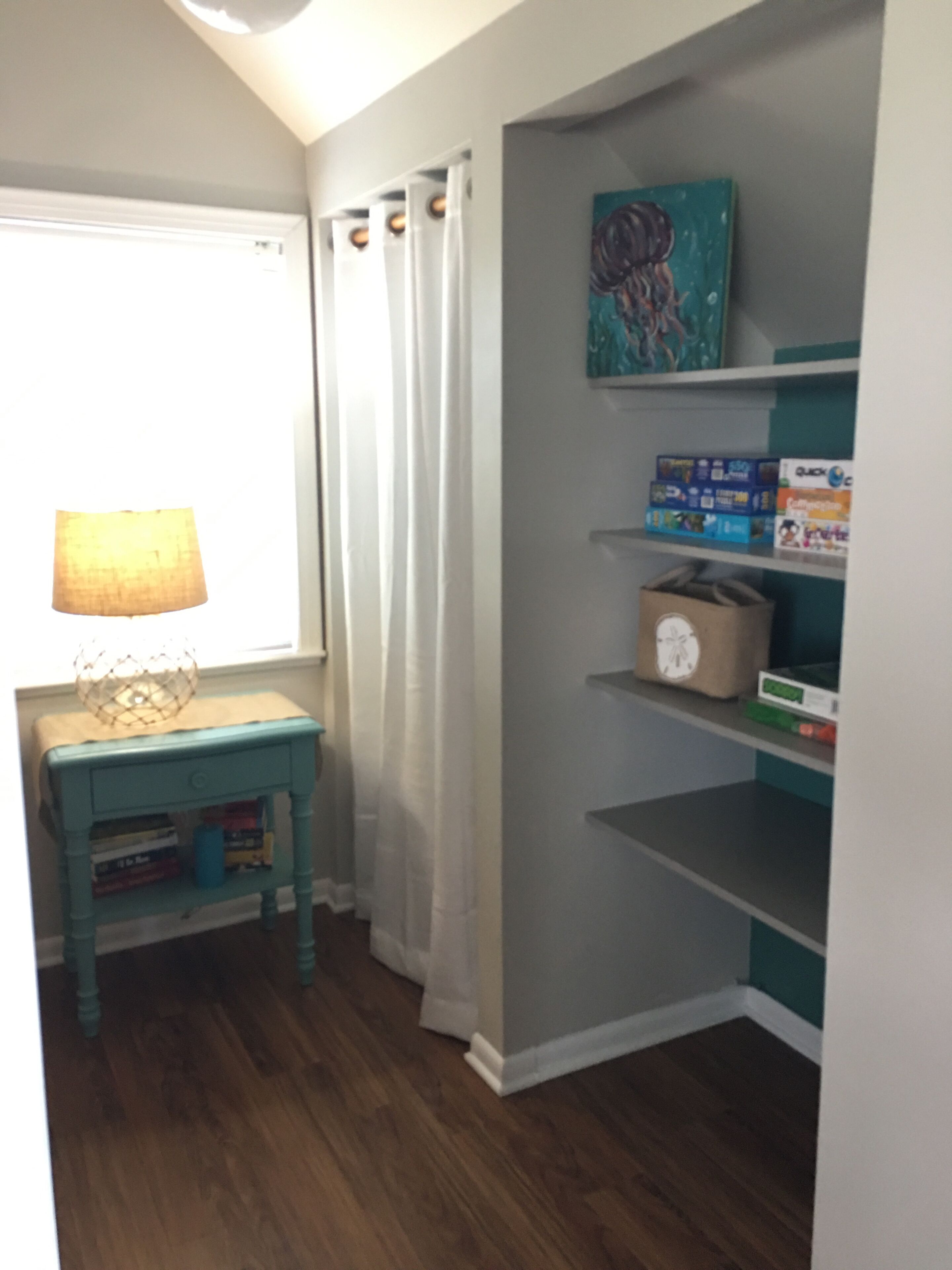 Une pièce avec une table de chevet de couleur turquoise, une lampe, une étagère avec des livres et une horloge, ainsi qu’une fenêtre avec des rideaux.