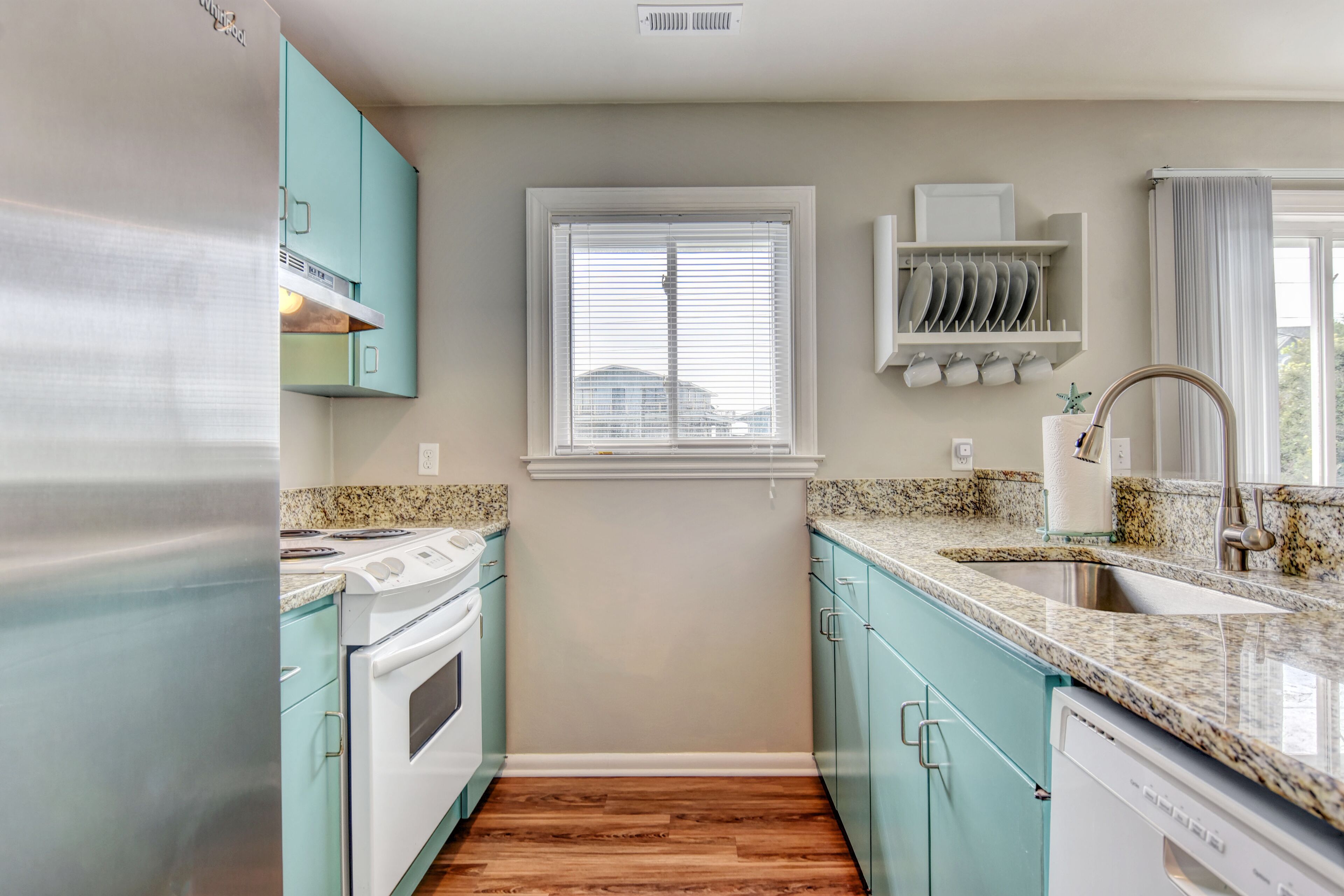 A kitchen with teal cabinets, a stainless steel refrigerator, a built-in oven, a granite countertop, and a window with blinds.