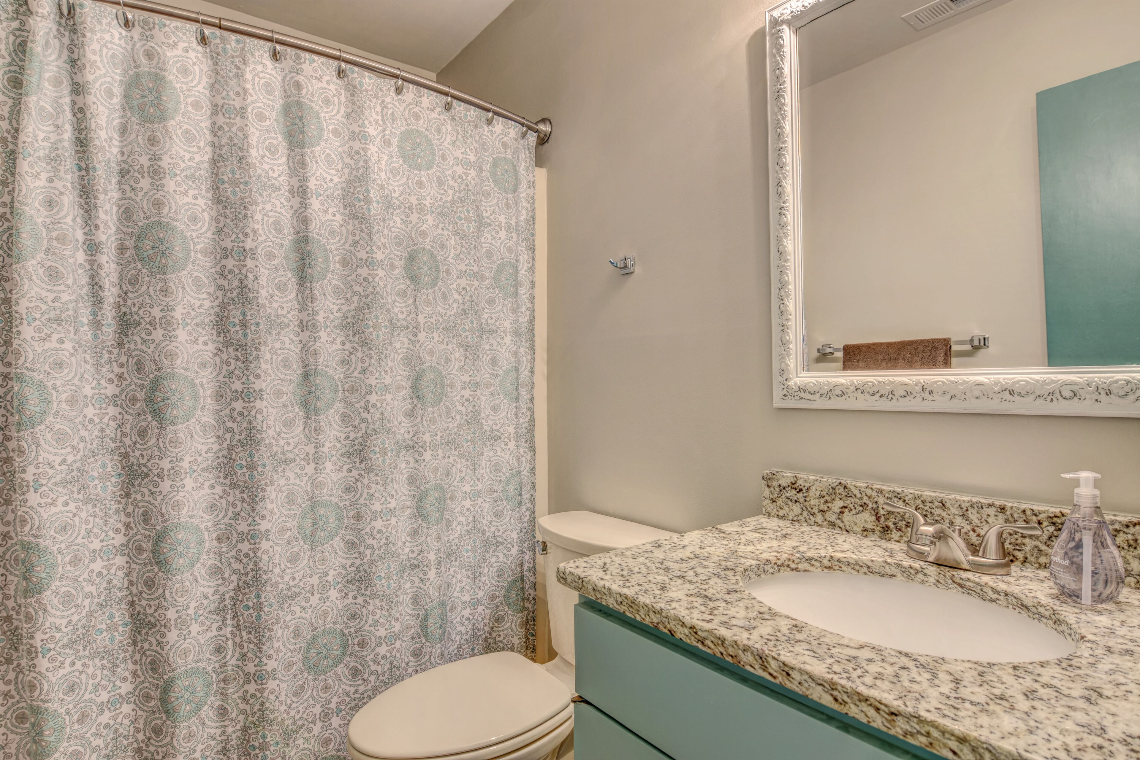 A bathroom with a patterned shower curtain, a toilet, a sink with a soap dispenser, and a mirror above the sink.