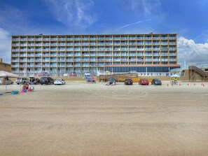 A large hotel with multiple floors and balconies, a parking lot with several cars, and a sandy ground area. - Oceanfront Condo | Hidden Treasure @ Pirates Cove (Daytona Beach)