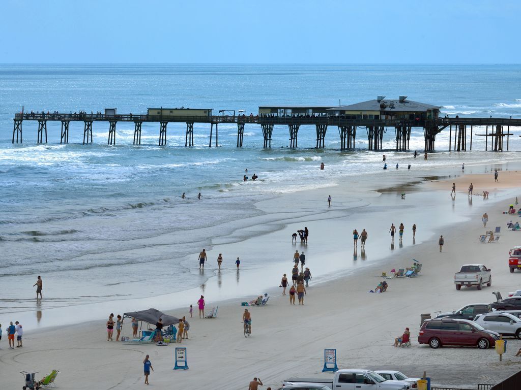 A beach with a pier extending into the ocean, people walking and relaxing on the sand, and several beach chairs and umbrellas.