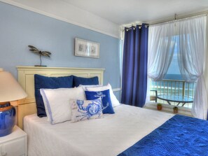 A bedroom with a bed, a nightstand, a lamp, a framed picture, and a view of the sea through a window with curtains. - Oceanfront Condo | Hidden Treasure @ Pirates Cove (Daytona Beach)