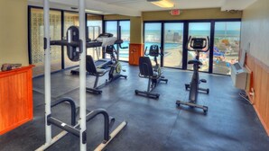 Uma academia com diversos equipamentos de exercício, grandes janelas com vista para a praia e um sinal de saída.