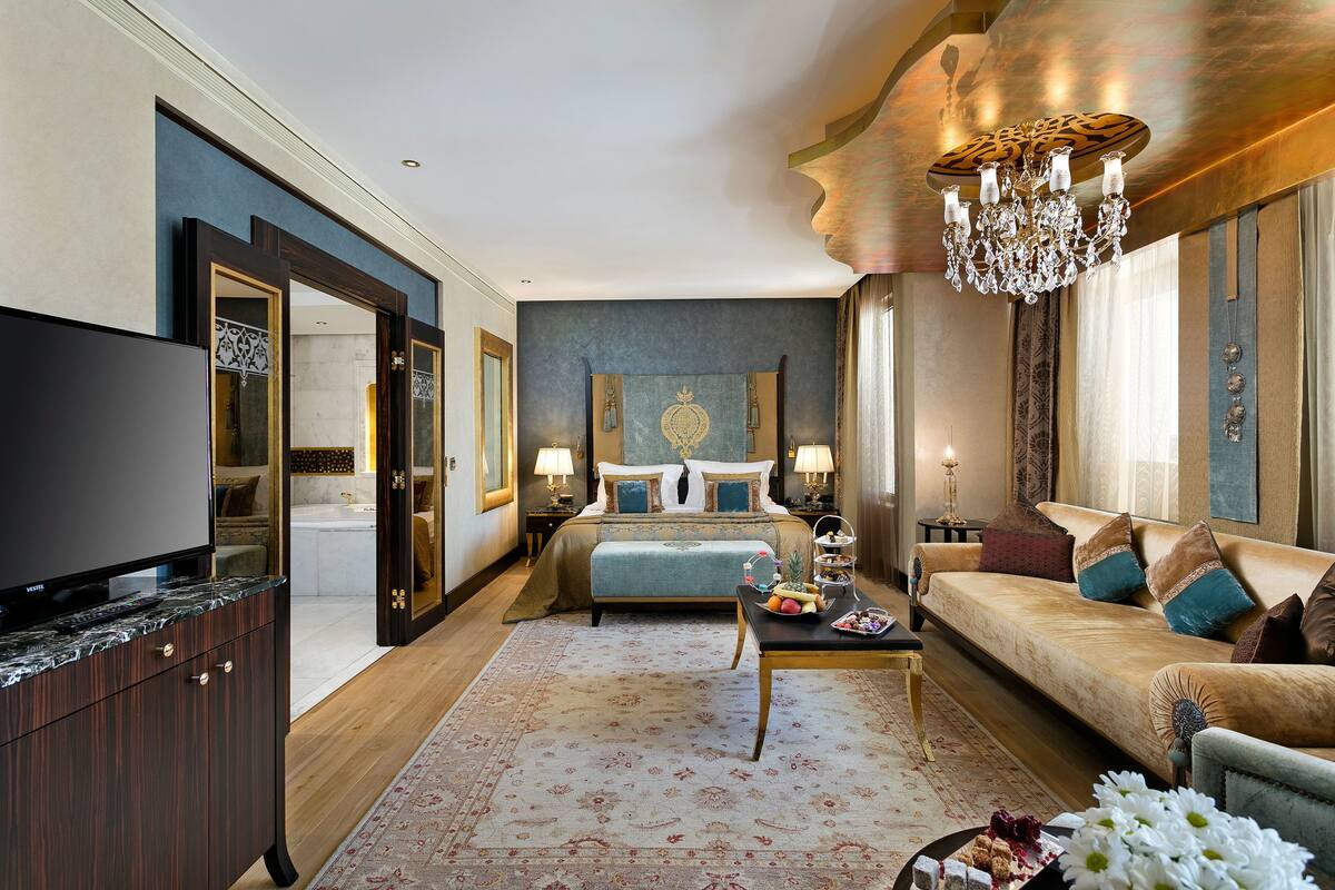 A spacious bedroom with a large bed, a sitting area with a sofa, a television, and a chandelier.