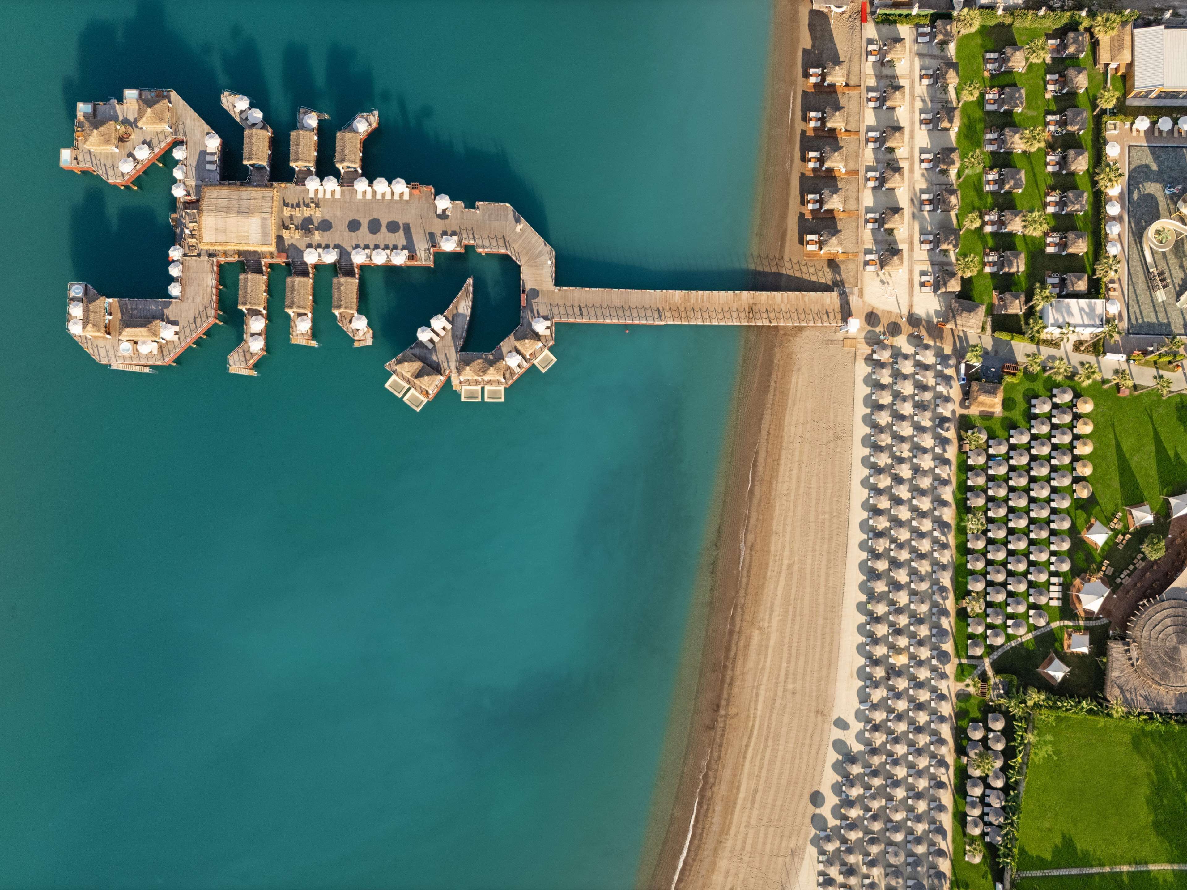 An aerial view of a coastal resort with a pier extending into the sea, a sandy beach, and a well-maintained lawn area with numerous umbrellas.
