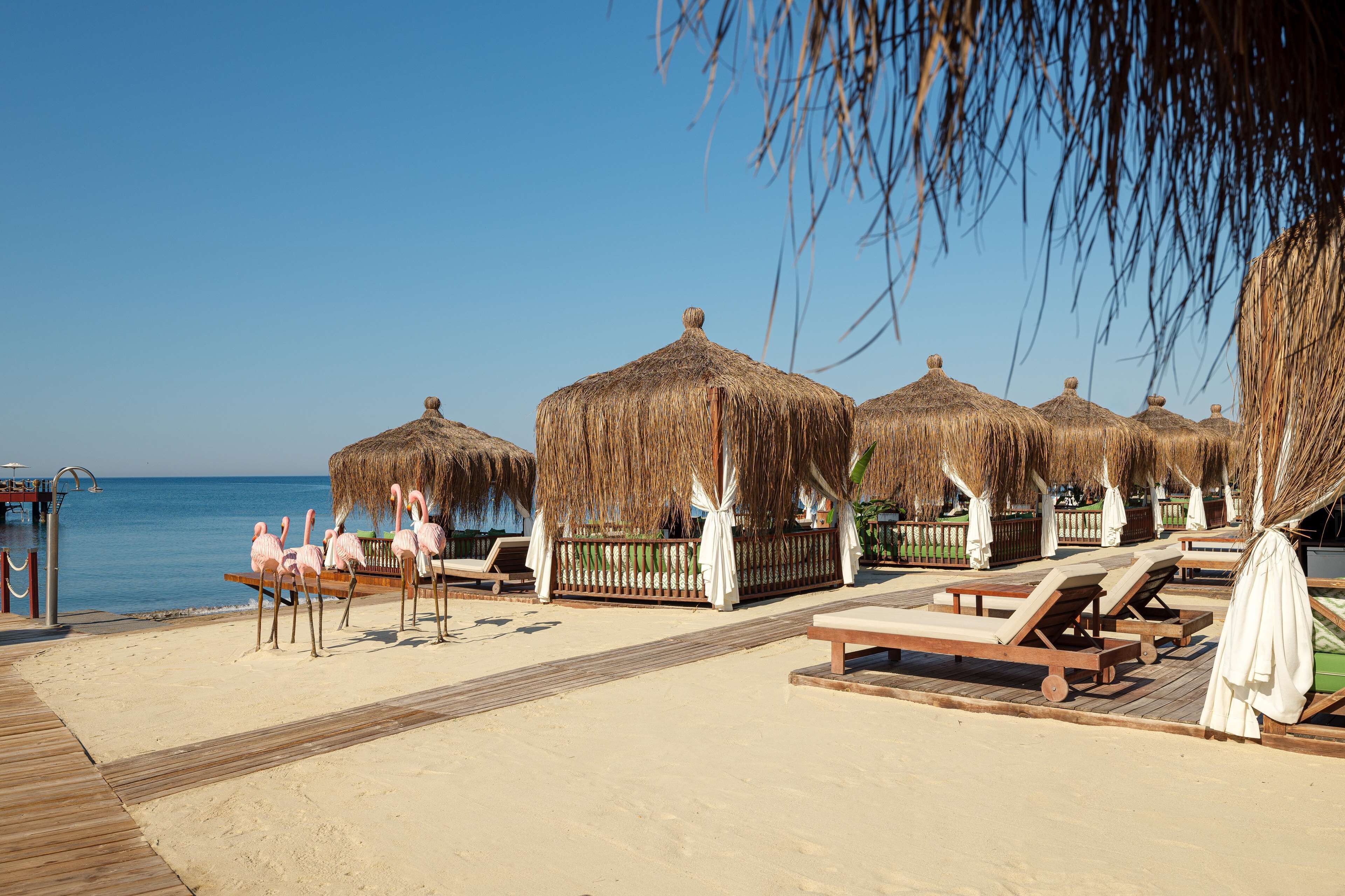 Una playa con sombrillas de paja, sillones de madera y un paseo de madera.