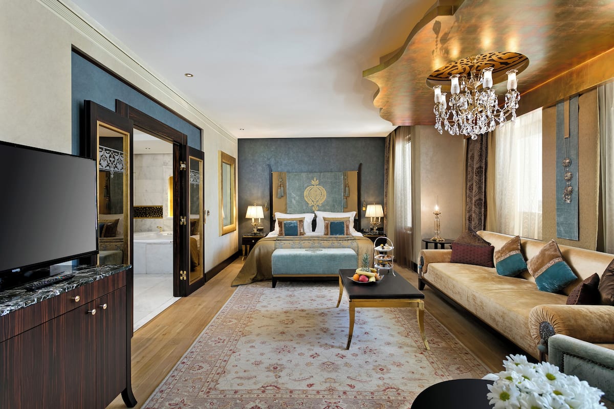 A spacious bedroom with a large bed, a sitting area with a sofa, a television, and a chandelier.