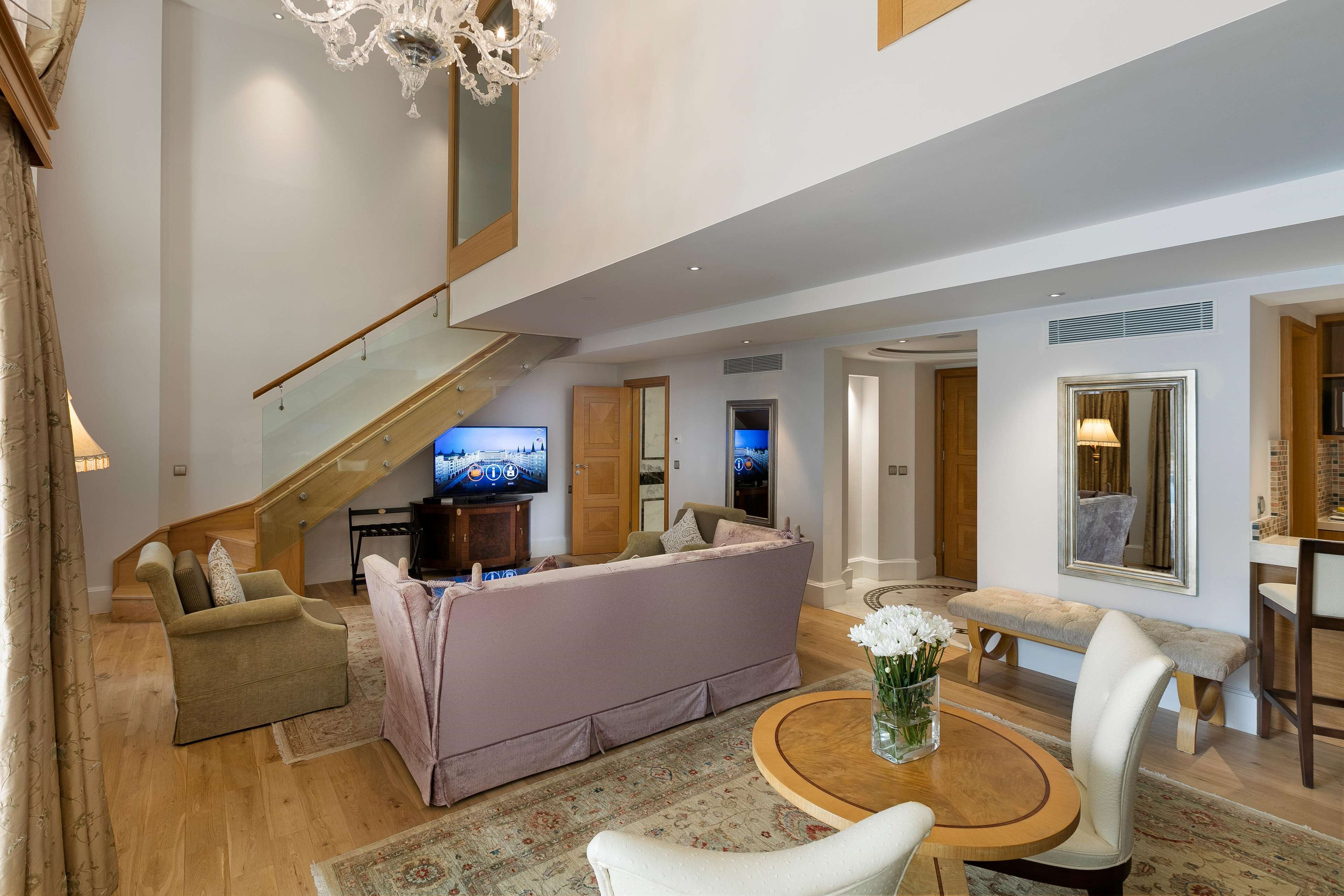 A spacious living area with a high ceiling, a staircase, a large flat-screen TV, a sofa, a bench, and a dining table.