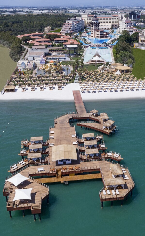 An aerial view of a resort with a large pool, multiple buildings, and a pier extending into the water. - Mardan Palace - All inclusive (Antalya)
