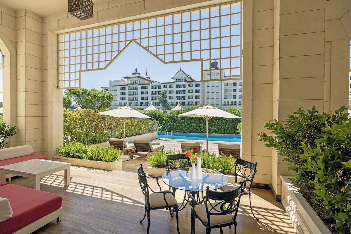 A sunlit outdoor seating area with a pool view, featuring a round table, chairs, and umbrellas.